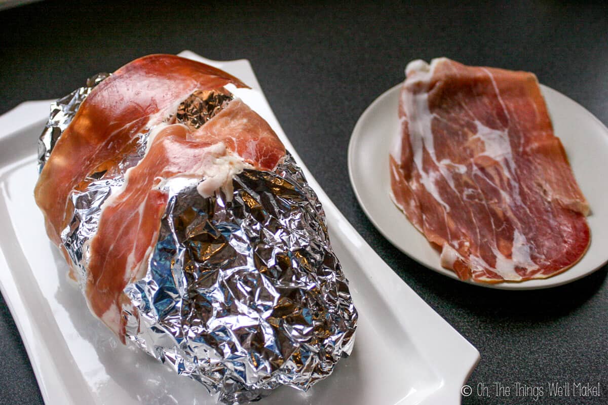 The foil covered face with two slices of prosciutto ham on it, on a white platter with a plate full of prosciutto ham on the side.