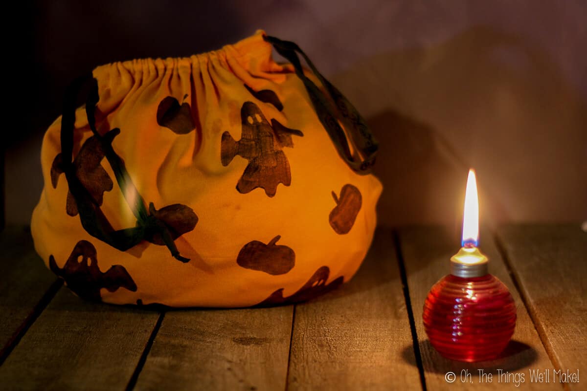 A homemade orange trick-or-treat fabric bag decorated with ghosts and bats laid on the left side in the dark with a small lighted red oil lamp on the other side.