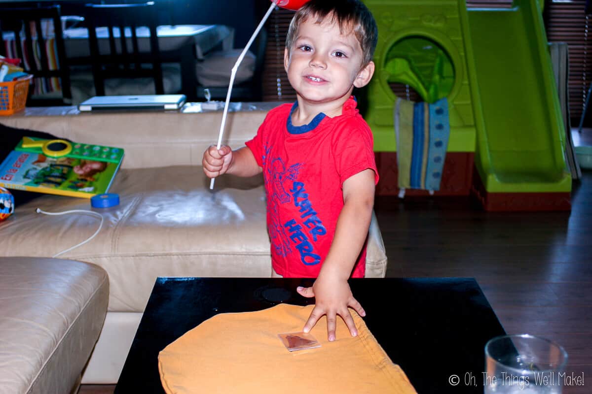 A young boy wearing a red shirt and holding a white balloon stick  eagerly smiling for the camera as he places a hand on an unmade trick-or-treat bag on the table.