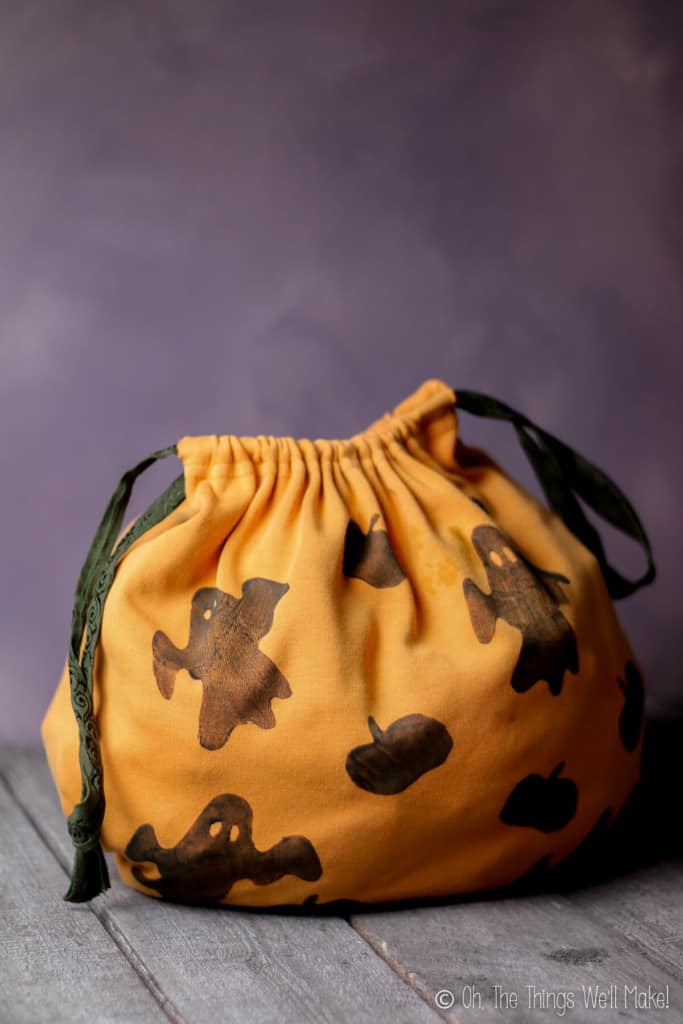 An orange handmade trick-or-treat fabric bag decorated with ghosts and bats stamps placed on a grey wood surface and grey photo backdrop.