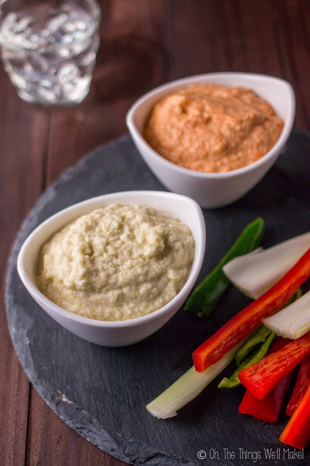 Two bowls of zucchini hummus on a black slate platter with cut veggies. Bottom bowl is filled with plain zucchini hummus while the bowl on top is orange zucchini hummus flavored with roasted red peppers.