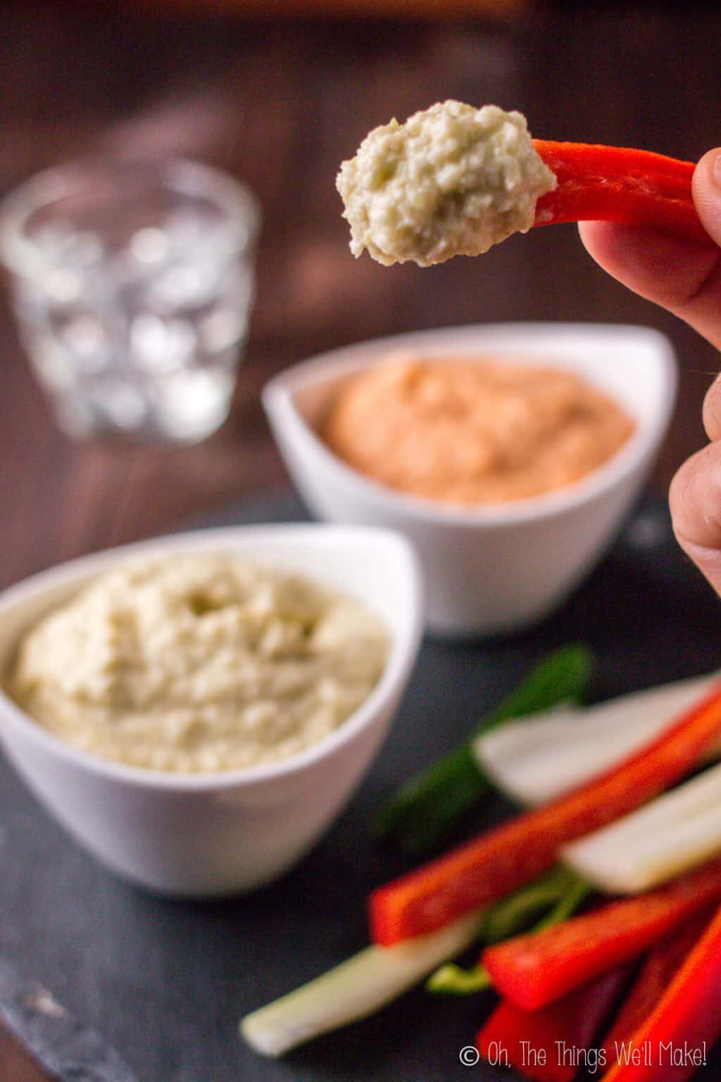 Close-up of a hand holding a red pepper strip topped with zucchini hummus with two bowls of zucchini hummus in the background.