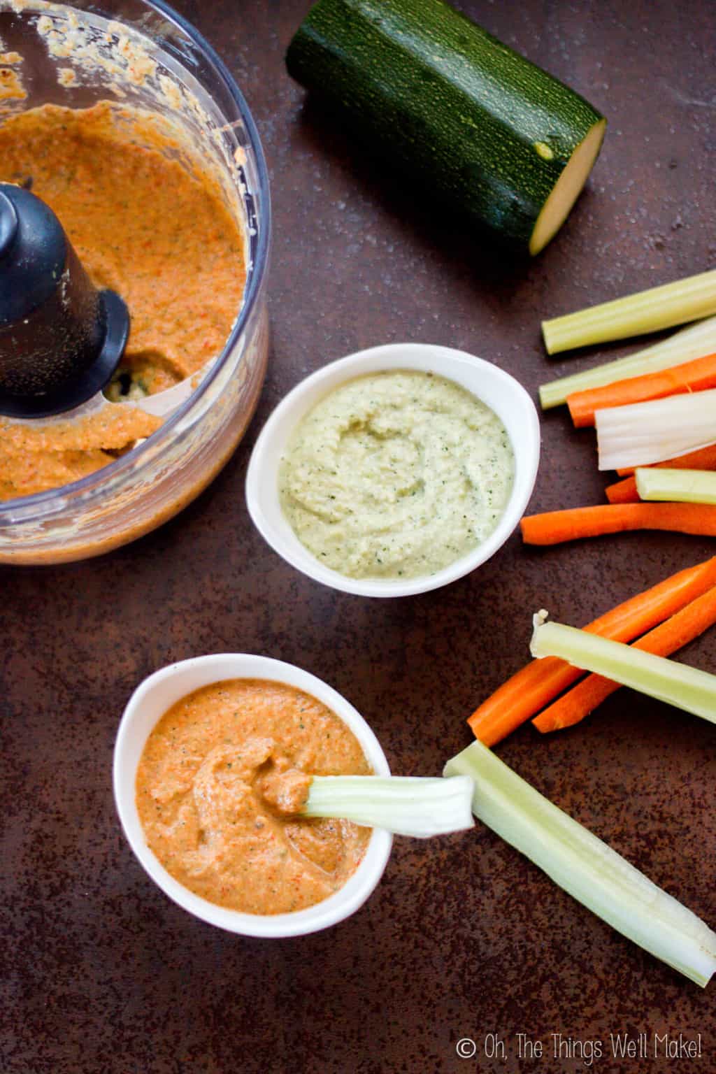 Top view of a food processor filled with zucchini hummus and roasted red peppers beside two bowls of zucchini hummus and cut up carrots, celery, and cucumber. The bowl in the bottom is filled with zucchini hummus flavored with roasted red pepper with a celery stick in it. The bowl on top is plain zucchini hummus.