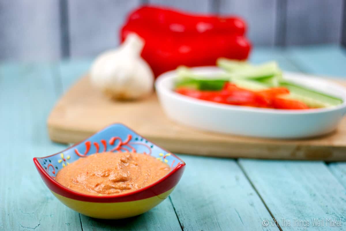 Close up of a bowl of roasted red pepper zucchini hummus in a bowl in front of some raw cut veggies.