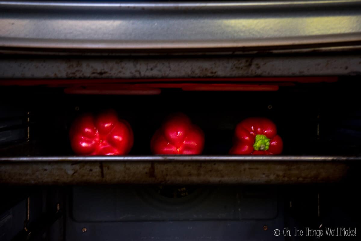 Three red peppers on a baking sheet under a broiler.