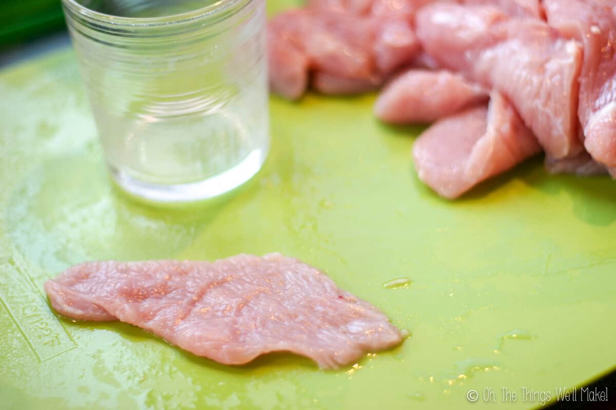 Close up of a pounded chicken breast fillet on a green chopping board with a glass beside it and several pieces of chicken fillet on the side.