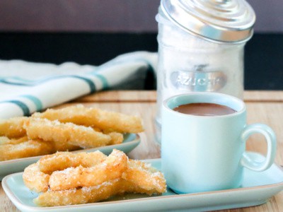 Overhead view of two plates of churros sprinkled with sugar with a cup filled with Spanish hot chocolate next to it.