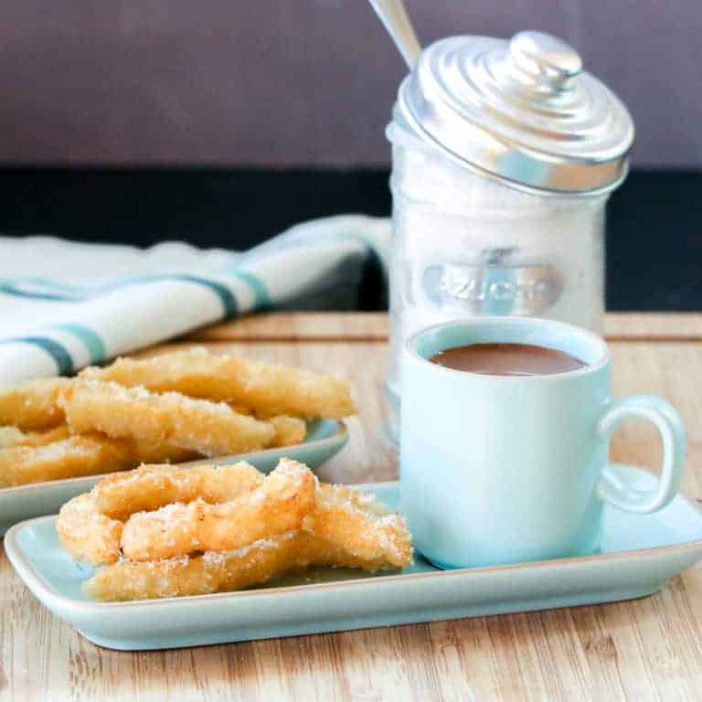 Overhead view of two plates of churros sprinkled with sugar with a cup filled with Spanish hot chocolate next to it.