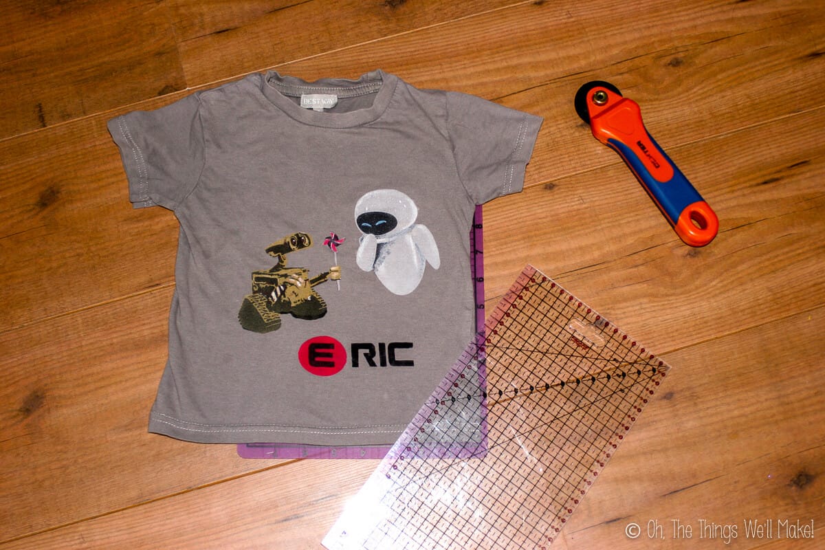 Overhead view of grey Wall-E t-shirt, a rotary cutter, and a plastic ruler mat, laid on a wood surface.