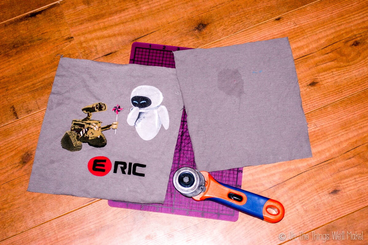 Overhead view of two squares of grey fabric cut from a t-shirt, laid on a purple cutting mat with a rotary cutter on the bottom. One square has a Wall-E design on it and one is plain.