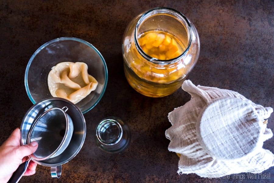 homemade vinegar in a jar with the mother in a bowl and a funnel and strainer being placed on a bottle to strain the vinegar.