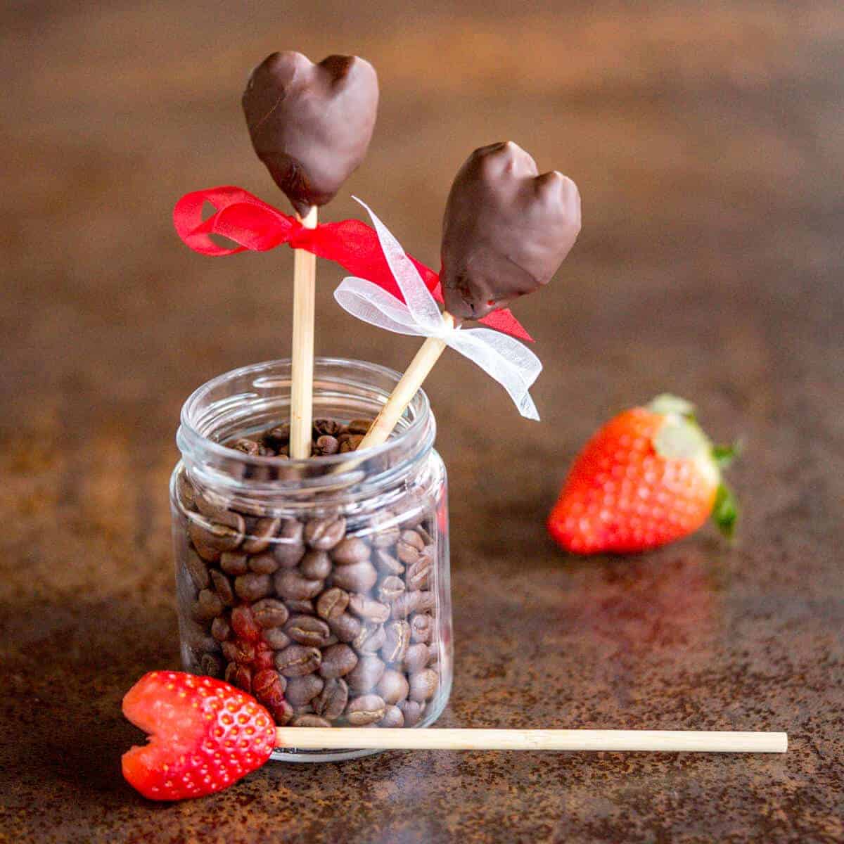 two chocolate-covered strawberry hearts in a jar next to a whole strawberry and a heart-shaped strawberry on a skewer ready for dipping in chocolate.
