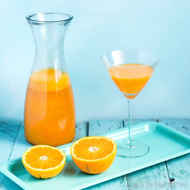 Oranges and a martini glass filled with agua de Valencia on a tray in front of a carafe of more of the orange-based cocktail.