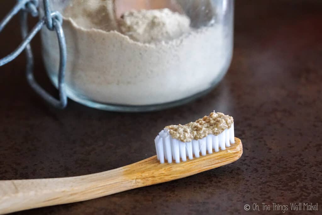 Closeup of tooth powder on a bamboo toothbrush. In the background theres a glass jar filled with the homemade tooth powder.