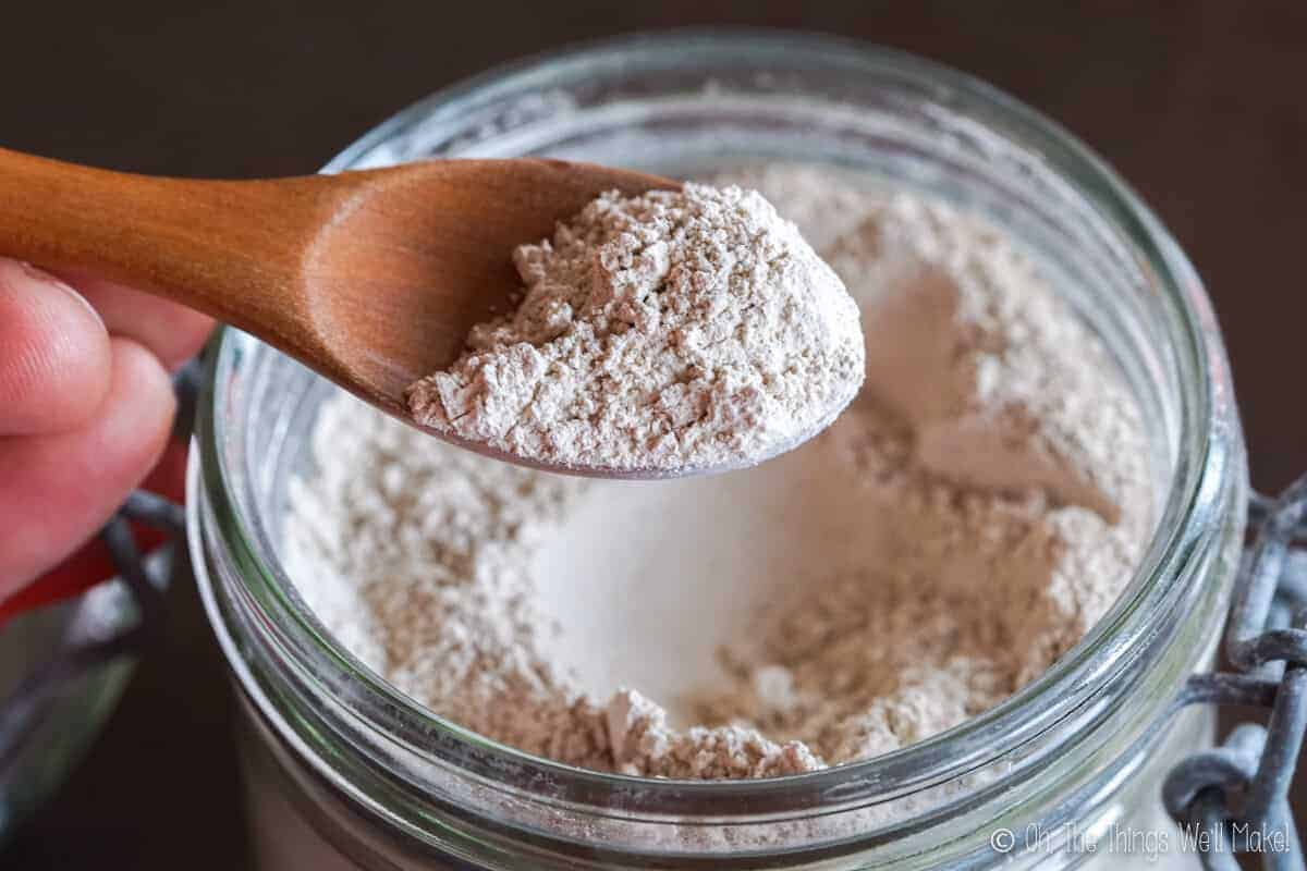 Close-up of a wooden spoon scooping bentonite clay powder from a glass jar, showing the fine, beige-colored natural clay often used in DIY tooth powder or skincare recipes.