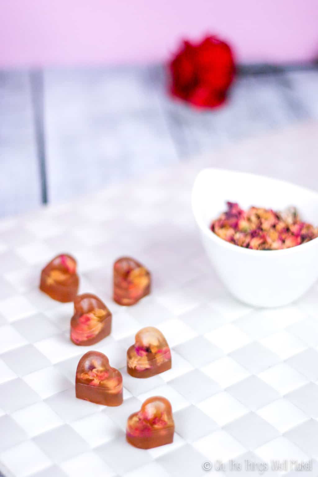 Close up of heart shaped gummy treats with real rose petals inside next to a bowl full of rose petals , placed on a white woven surface. A single red rose is at the background.