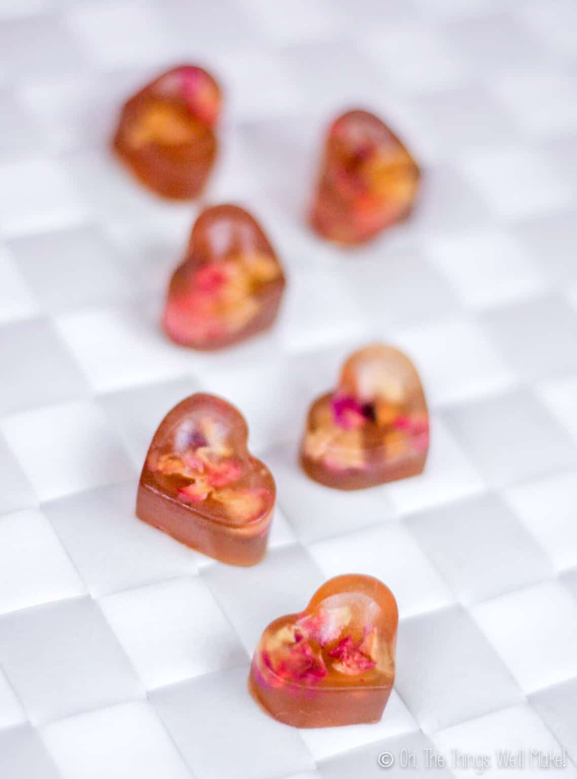 Close up of six heart shaped rose petal gummies on a white woven surface.