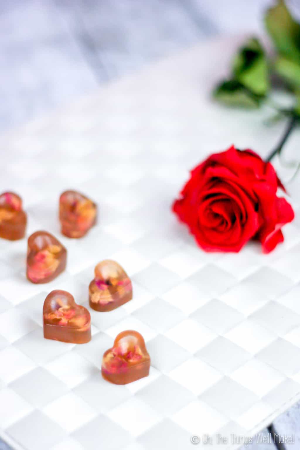 Close up of heart shaped gummy treats with real rose petals inside next to a red rose, placed on a white woven surface.