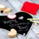 A homemade cloth chalkboard heart with "Happy Valentine's Day!" written on it. It's surrounded by homemade chalk and next to a folded chalkboard.