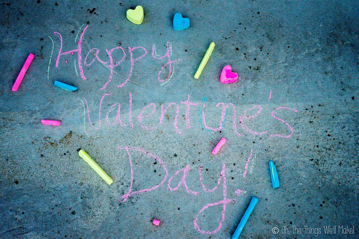 Overhead view of "happy Valentine's Day" written with homemade pink and blue chalk, with several pieces of heart shaped chalk and sidewalk chalk sticks laid around on the ground.