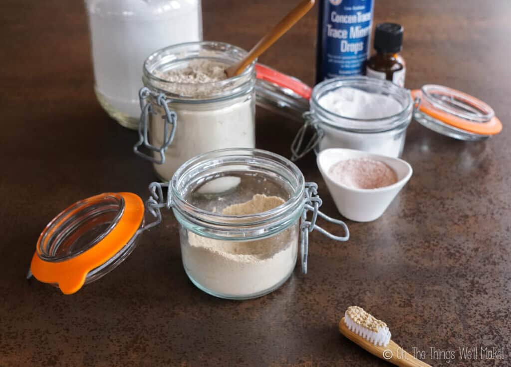 a jar of homemade tooth powder in front of jars of ingredients (salt, bentonite clay, baking soda, etc.