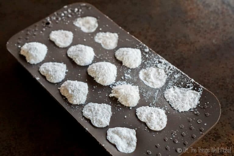 A silicone mold contains heart-shaped bath fizzies dusted with powdered sugar, resting on a dark surface.