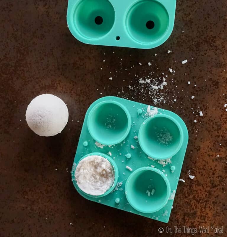 Green silicone mold with round cavities, one filled with powder, another empty, and a finished white bath fizzie resting beside the mold on a brown surface.