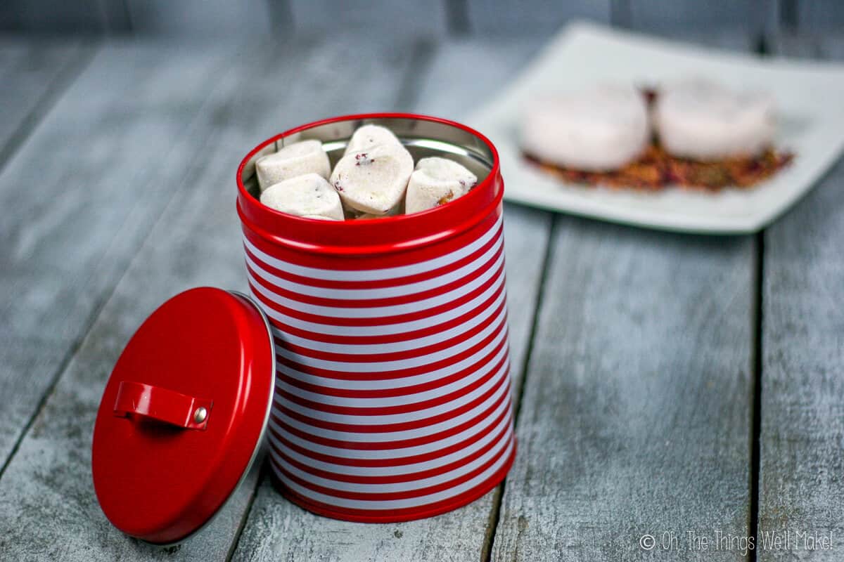 A red and white striped tin can filled with small rose petal bath bombs. Behind it, are two large bath bombs on a plate with rose petals.