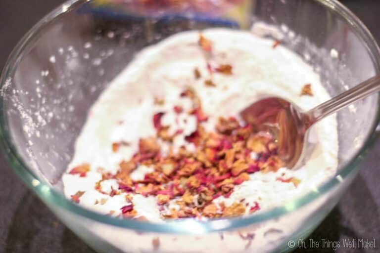 A glass bowl contains a white powder mixture for Bath Fizzies with dried rose petals, being stirred with a metal spoon.