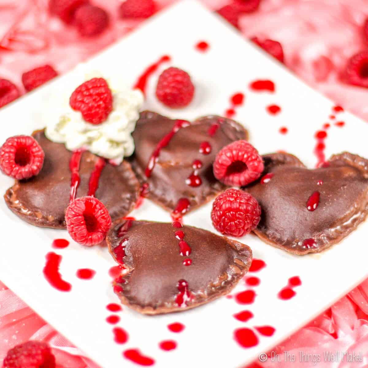 Four pieces of homemade chocolate heart shaped ravioli on a plate with raspberry puree, fresh raspberries and whipped cream