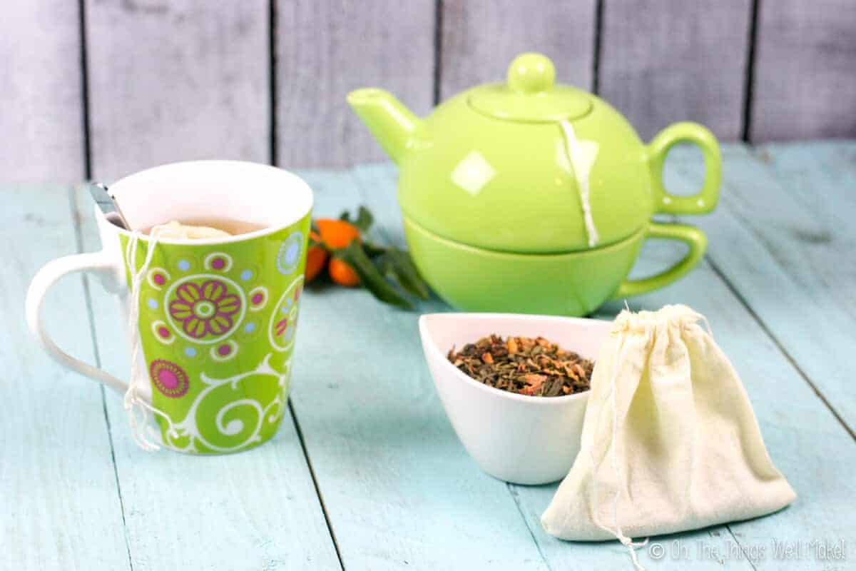 A cloth teabag in front of a small bowl with loose tea in it. In the background, there is a cup of tea and a teapot.