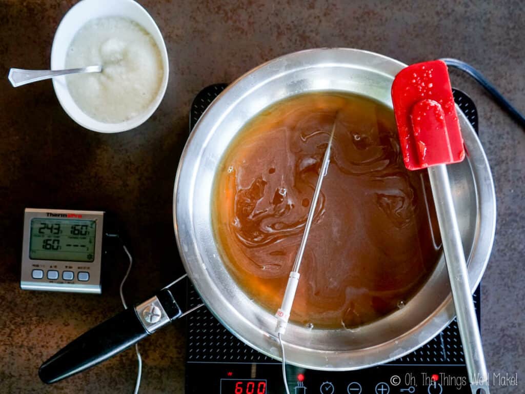 Overhead view of a honey and water mixture being heated in a pan, and with a thermometer probe inside it. Gelatin is hydrating in a white bowl above the pan.