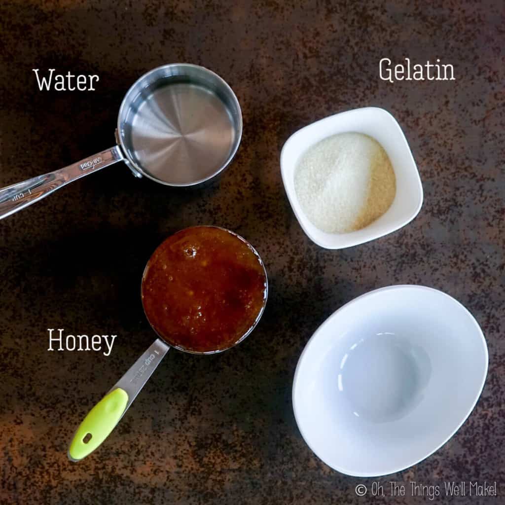 Overhead view of a cup of water, a cup of honey, some gelatin powder in a small bowl, and an empty bowl for mixing ingredients.