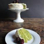 a slice of a nettle cake with white frosting, garnished with raspberries, in front of a cake on a stand.