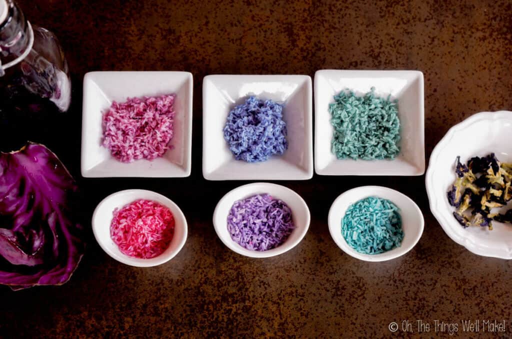 overhead view of 6 bowls of shredded coconut that has been colored with red cabbage and butterfly pea flowers. The two on the right are turquoise, the middle ones are blue and purple, and the ones on the left are pink. To the right of the bowls is a bowl of butterfly pea flowers, and on the left are some red cabbage leaves.