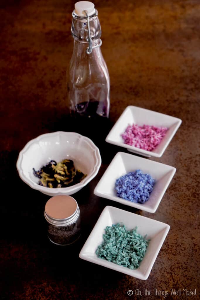 Overhead view of 3 small bowls of colored shredded coconut next to a bowl of butterfly pea flowers and a bottle of blue liquid obtained from the flowers. The top bowl has pink shredded coconut, the middle one has purple, and the bottom one has turquoise shredded coconut.