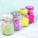 4 glass jars filled with 4 different types of colorful sprinkles made from shredded coconut. From left to right: green, purple, yellow, and bright pink.