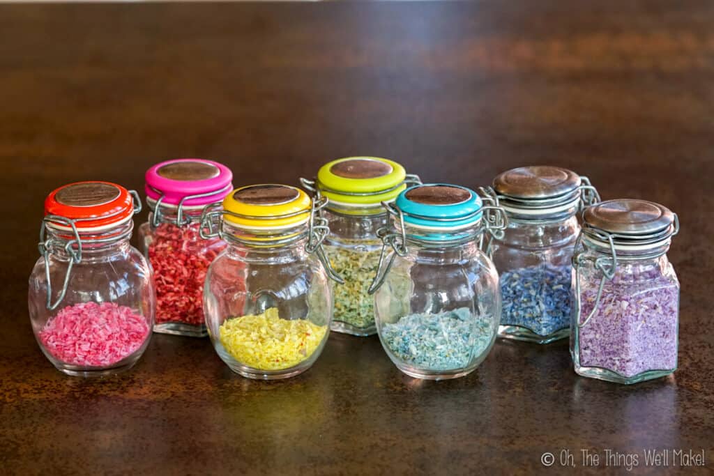 7 small glass jars filled with 7 different types of colorful sprinkles made from shredded coconut in a rainbow of colors