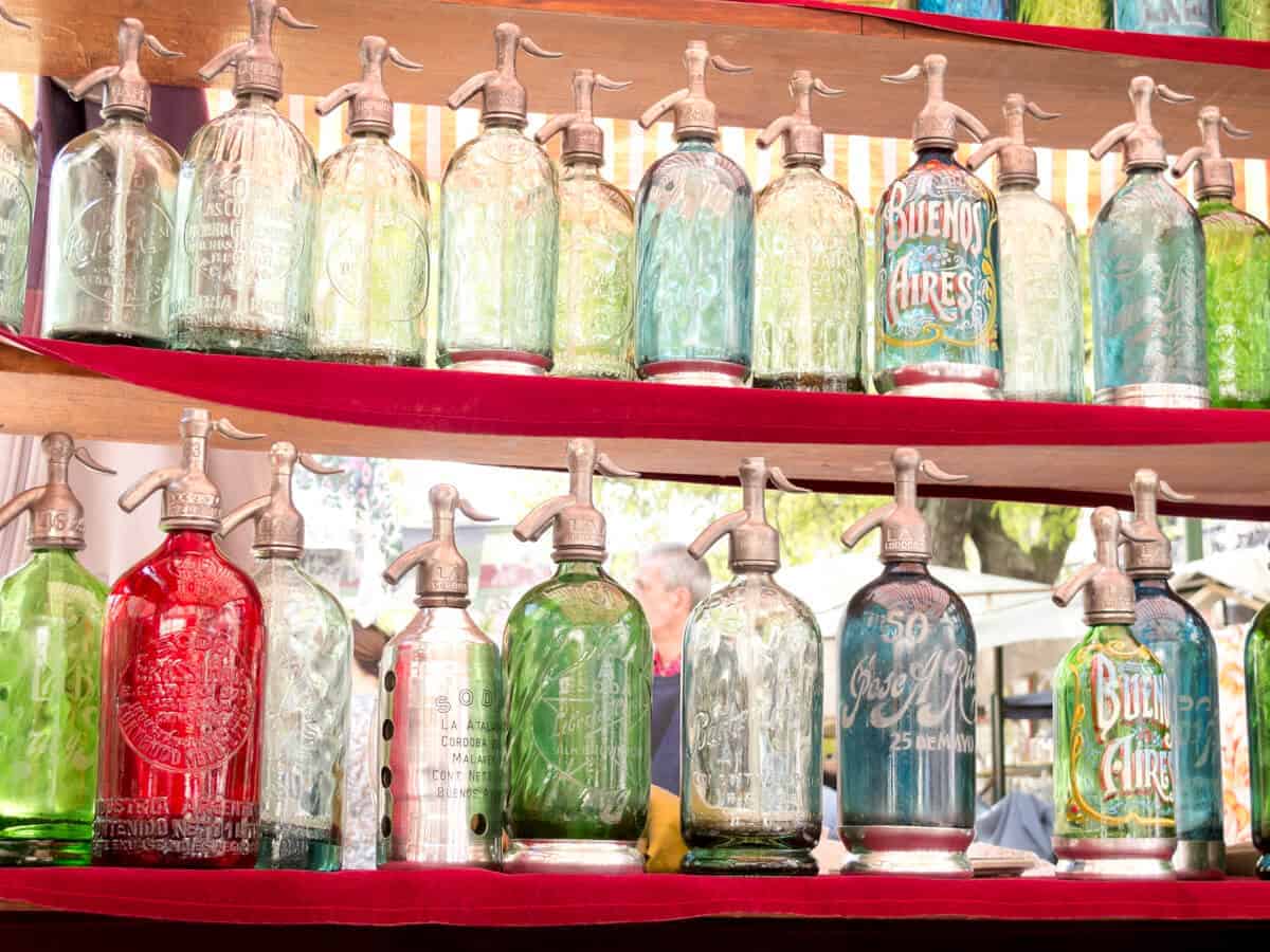 Two shelves filled with antique glass soda siphons in a variety of colors