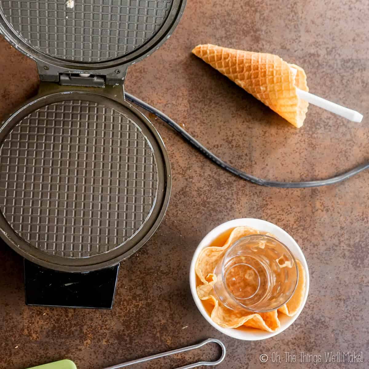 Overhead view of an open waffle cone iron, next to a homemade waffle cone and waffle bowl