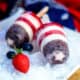 Closeup of two patriotic popsicles (red, white, and blue) on a plate with ice and a strawberry and a blueberry.