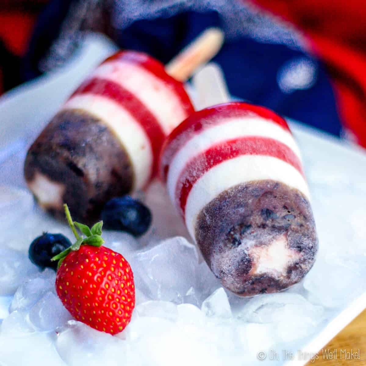 Closeup of two patriotic popsicles (red, white, and blue) on a plate with ice and a strawberry and a blueberry.