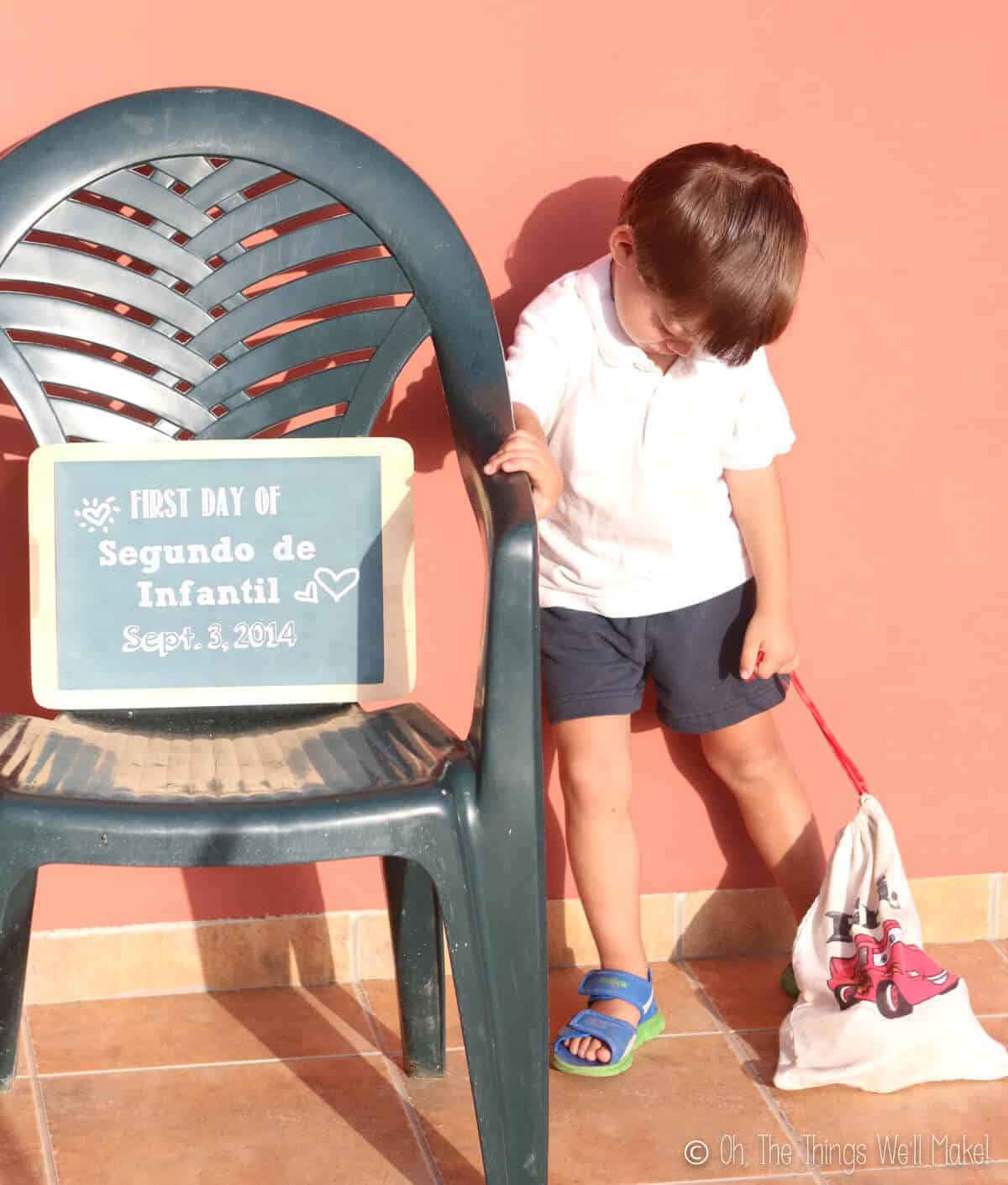 Upset young boy with his head down next to a sign made to memorialize the first day of school