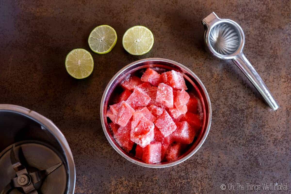 Overhead view of a bowl of frozen watermelon chunks, some limes, and a lime squeezer