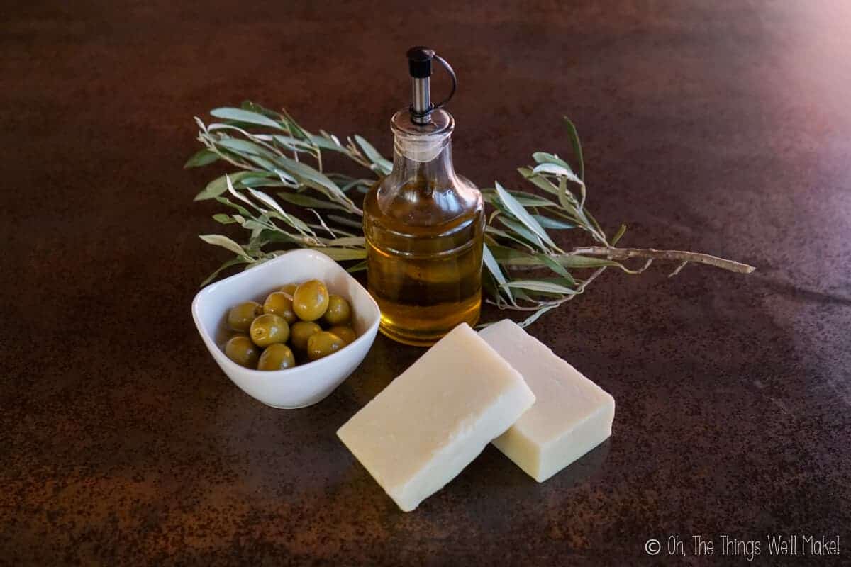 Olive oil in a bottle near a bowl of olives and 2 bars of Castile soap