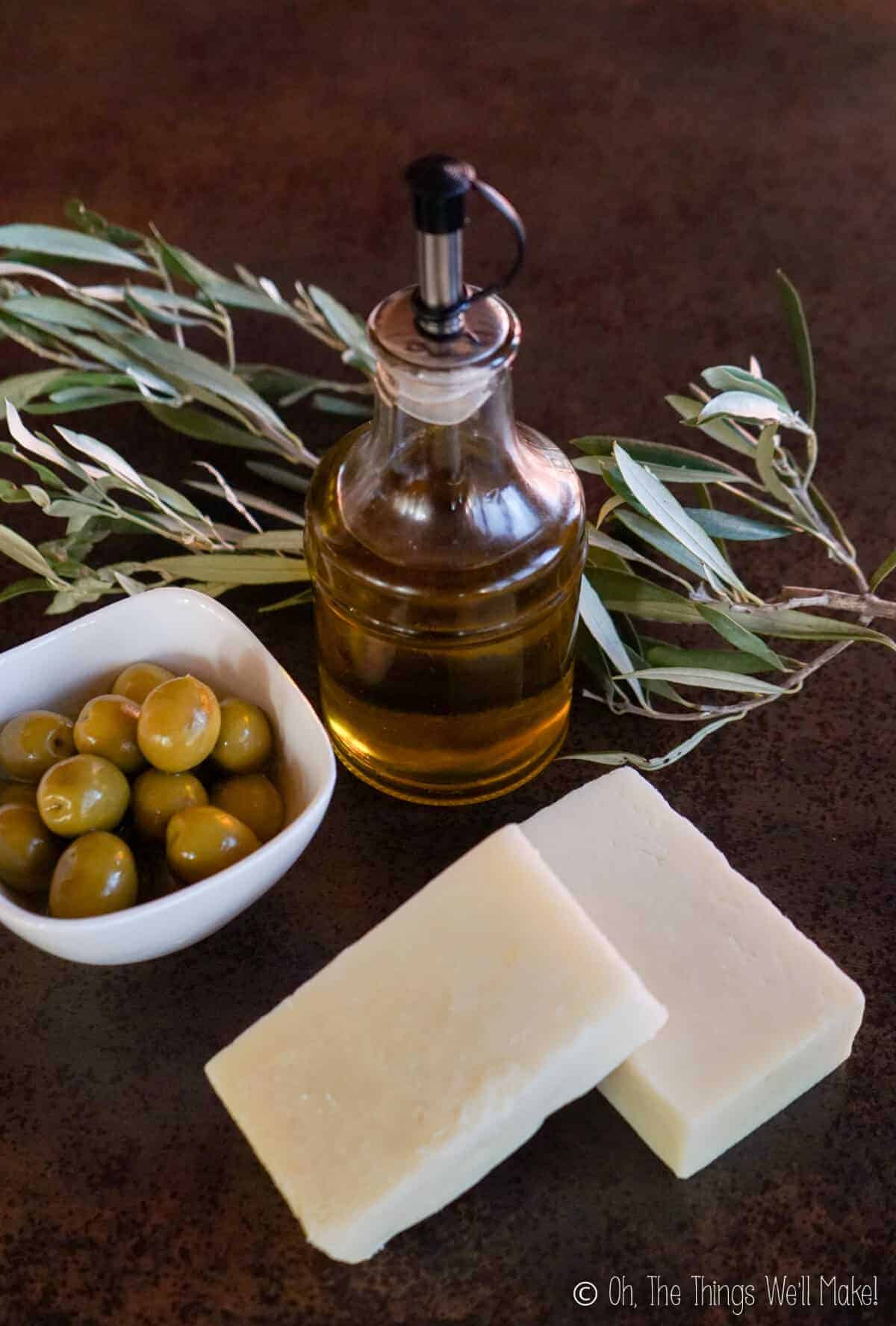 Olive oil in a bottle near a bowl of olives and 2 bars of Castile soap