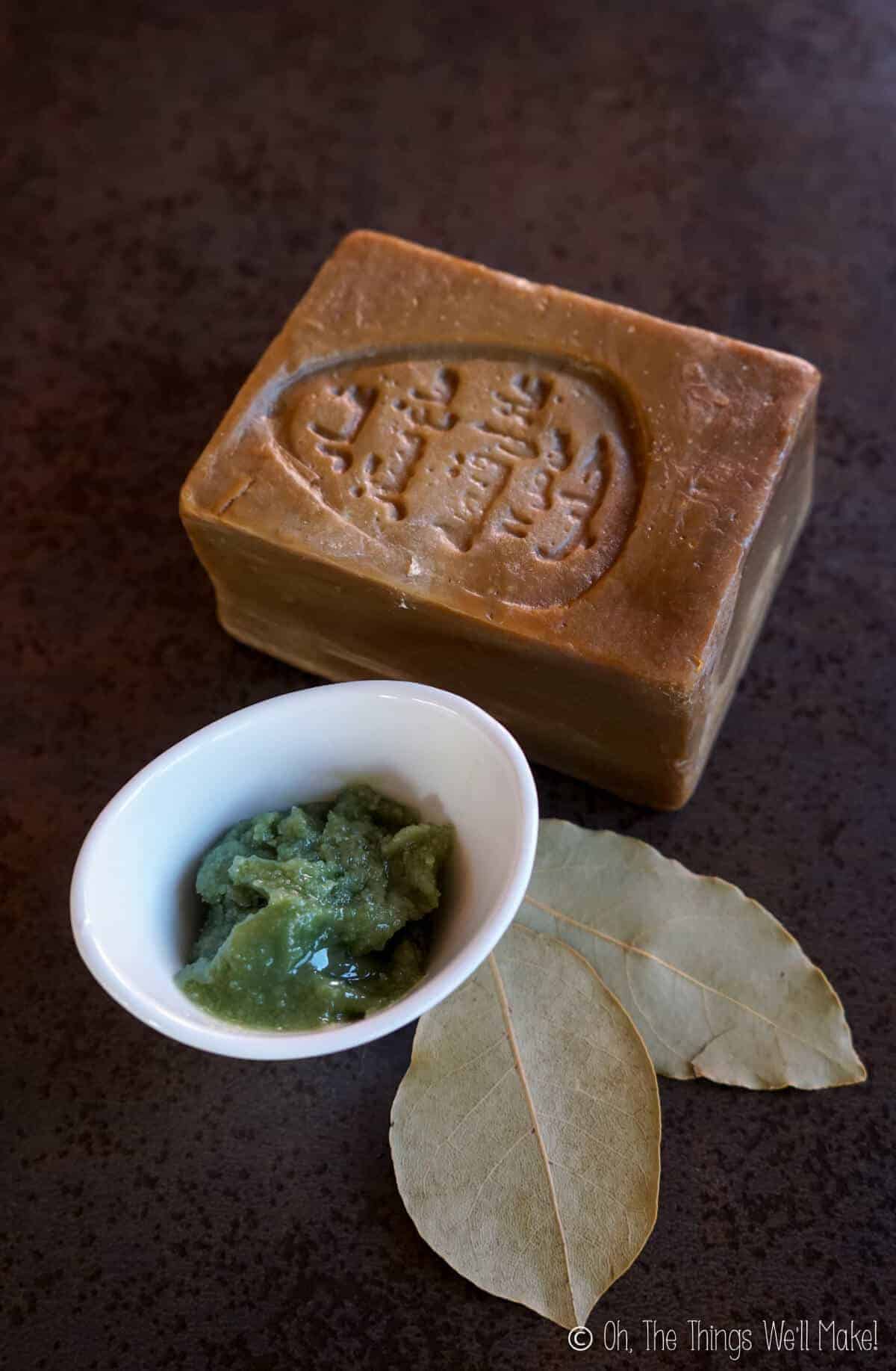 Overhead view of a small container with laurel berry oil next to some bay leaves and a bar of Aleppo soap