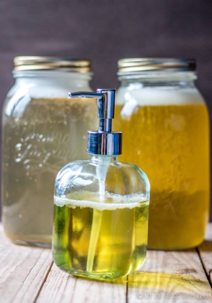 a pump bottle filled with liquid soap in front of 2 jars of liquid soap. (The one on the left was made with coconut oil and is lighter in color. The one on the right, made with olive oil, is amber in color.)