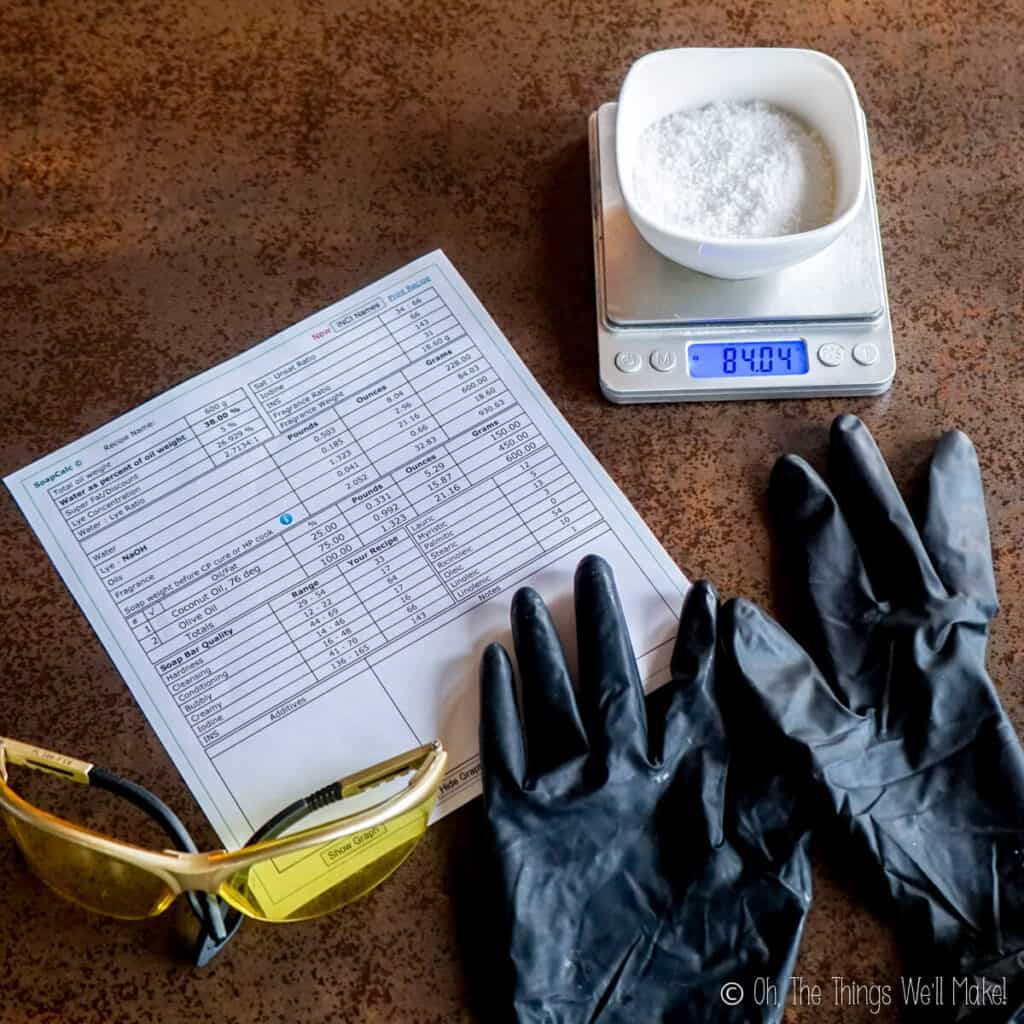 Overhead view of.a soap recipe as printed from a lye calculator. Next to the recipe are some gloves, safety glasses, and a bowl with lye in it. 