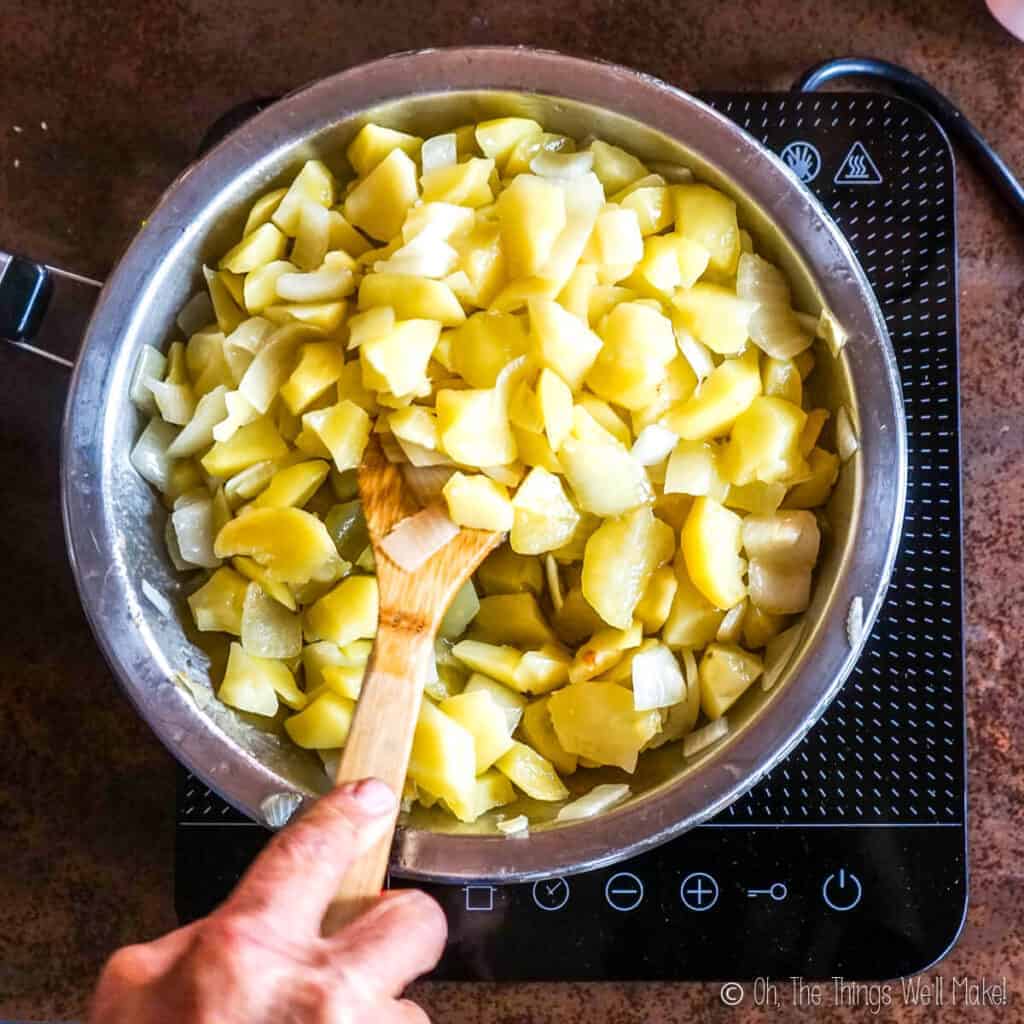 Overhead view of potatoes and onions in a frying pan with a wooden spatula being used to stir them